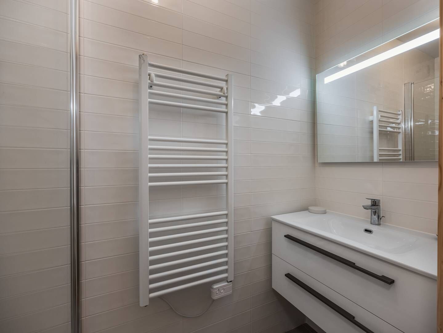 A modern bathroom with a glass shower enclosure, a white tiled wall, a white sink with a mirror, and a towel rack.