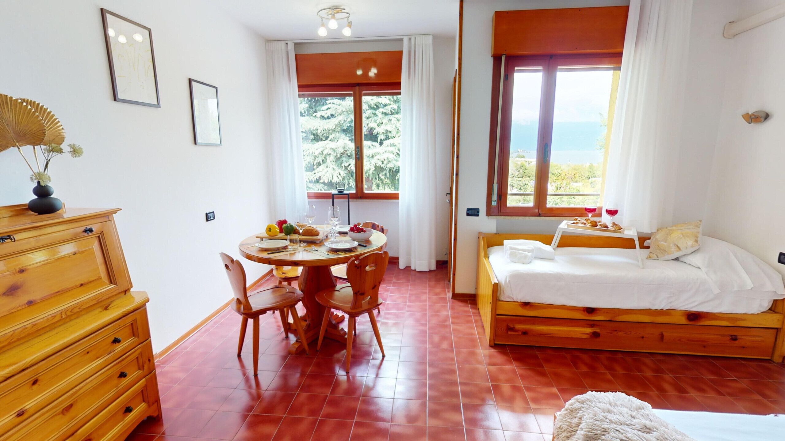 Una habitación con una cama de madera, una mesa de comedor con sillas, un aparador y una ventana que da a vistas de árboles y el mar.