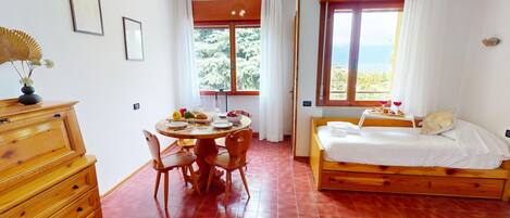 Una habitación con una cama de madera, una mesa de comedor con sillas, un aparador y una ventana que da a vistas de árboles y el mar.