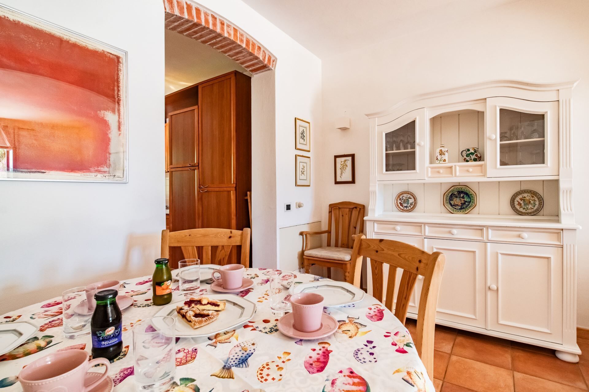Un comedor con una mesa dispuesta para dos personas, con mantel floral, sillas de madera y un mueble blanco con puertas de vidrio que exhibe platos decorativos.