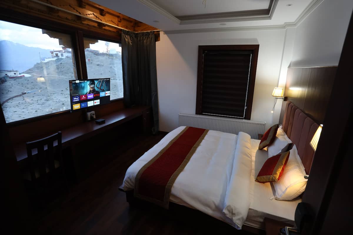 A hotel room with a bed, a desk with a computer, a window with a view of a mountainous landscape, and a lamp on the wall.