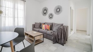 A modern living room with a grey sofa, a coffee table, and a dining table with chairs. There are decorative wall mirrors and a plant on the windowsill.