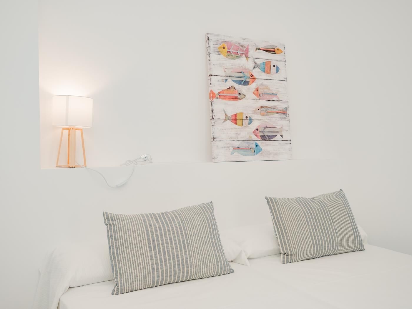 Une chambre à coucher avec un lit blanc, deux coussins à rayures, une lampe fixée au mur et un tableau coloré représentant des poissons.