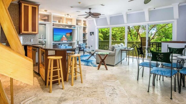 A spacious living area with a kitchen, dining area, and large windows offering a view of greenery. - Serenity Retreat- 2/2 Townhouse, Dock & Pool (Key West)