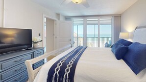 A hotel room with a bed, a television, a ceiling fan, and a balcony with a view of the ocean.