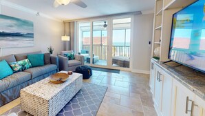 A spacious living room with a large television, a ceiling fan, and a balcony with a view.