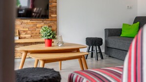 A modern living room with a wooden coffee table, a grey sofa with green cushions, a black ottoman, and a flat-screen TV mounted on a wooden wall. - Modern apartment with woodstove & large terrace (Leogang)