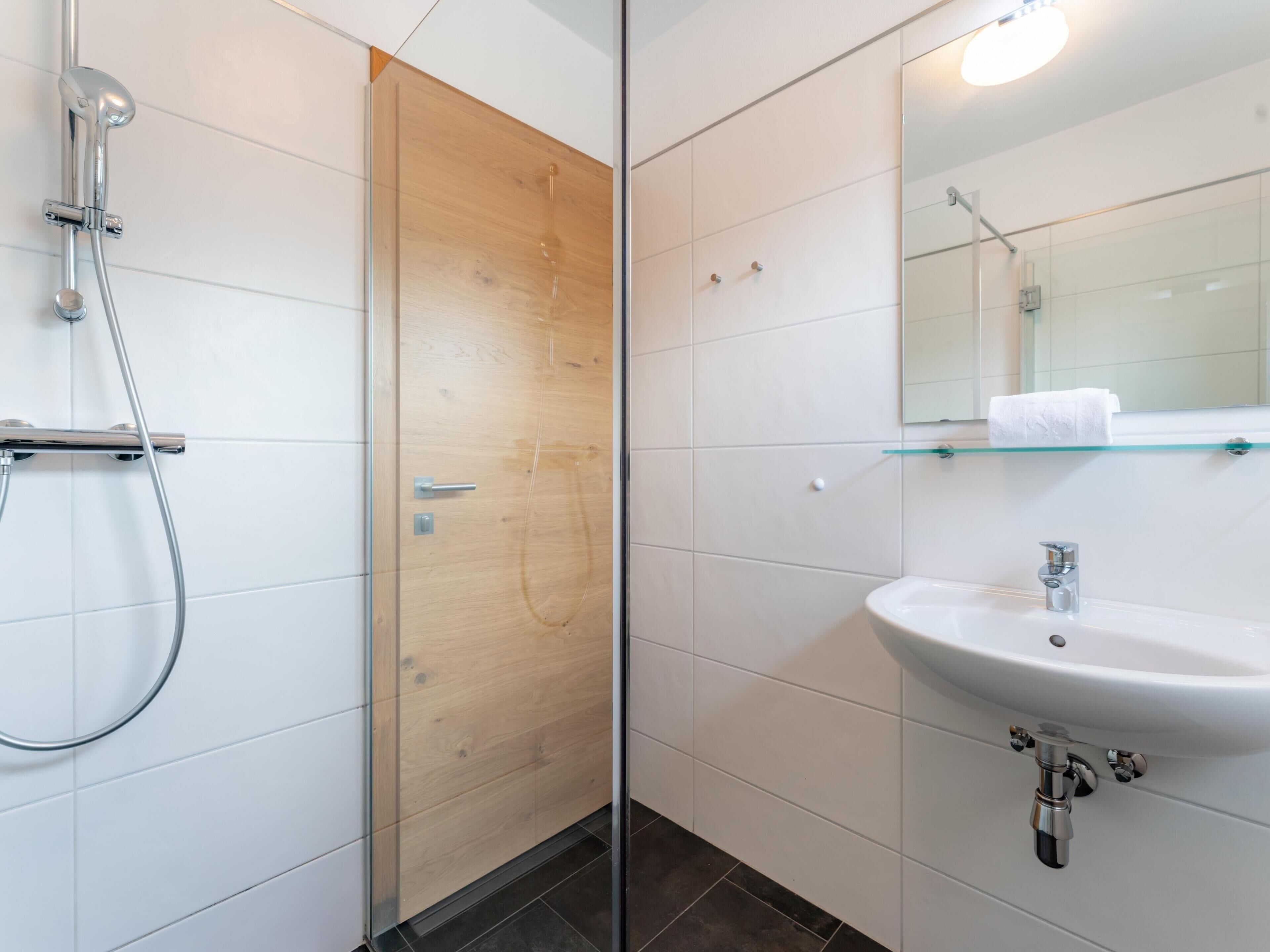 Een moderne badkamer met een douchecabine van glas, een houten deur, witte betegelde wanden, een glazen plankje met een handdoek, en een witte wastafel met een spiegel erboven.