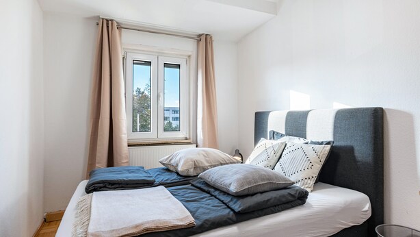 A bedroom with a bed, a window with curtains, a radiator, and a metal basket. - Holiday Apartment 'Ludwigsburg' with Wi-Fi (Ludwigsburg)