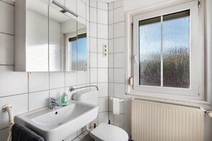 Une salle de bain avec un lavabo blanc, un miroir et des toilettes, une fenêtre donnant sur des arbres et un radiateur blanc.