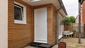 Fachada de madera con una puerta blanca, un pequeño banco y una planta en maceta.