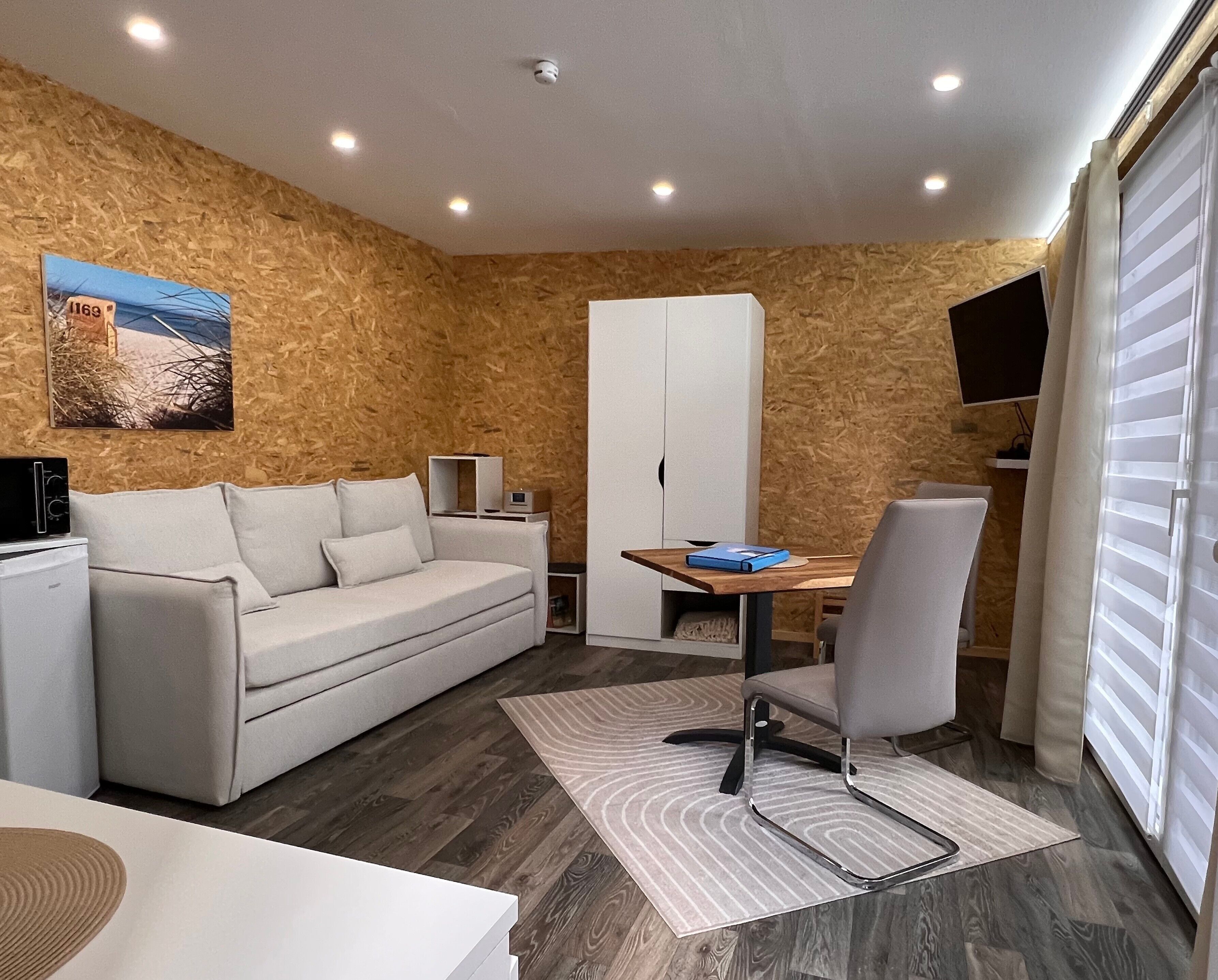A modern room with a sofa, a dining table, a chair, a TV, and a window with blinds.