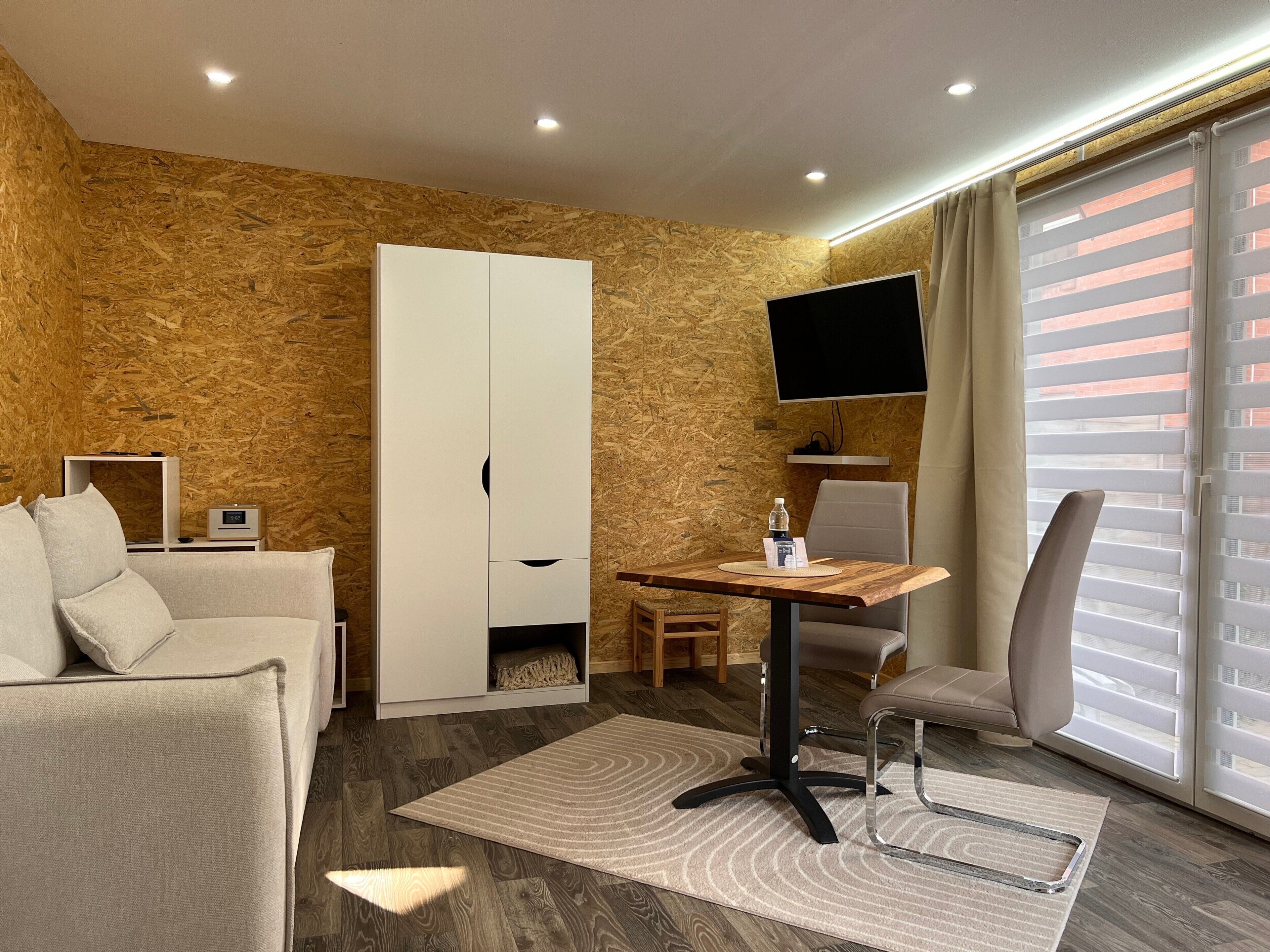 A modern hotel room with a wooden wall, a sofa, a TV, a desk with a chair, and a window with blinds.