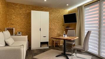 A modern hotel room with a wooden wall, a sofa, a TV, a desk with a chair, and a window with blinds.