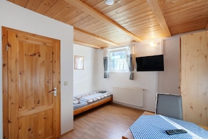 Una habitación con techo de madera, una cama, una mesa con mantel a cuadros, una silla, un televisor montado en la pared y una ventana con cortinas.