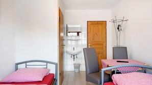 A small, clean room with a bed, a chair, a table, a sink, and a coat rack. - Knödelhof 'Room 3' with Balcony – Guesthouse near Munich (Putzbrunn)