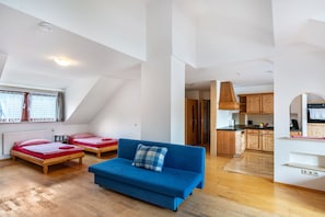 Un appartement de style « loft » comprenant un canapé bleu, deux lits simples, un coin cuisine et une table à manger.