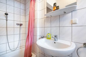 A bathroom with a shower, a sink, a red and white checkered shower curtain, and a shelf with toiletries.
