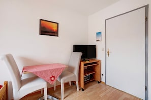 Una habitación con un sofá blanco, una mesita con mantel rojo, un televisor sobre un mueble de madera y una puerta con picaporte de latón.