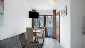Ein Zimmer mit einer Couch, einem an der Wand montierten Fernseher, einem Essbereich mit Stühlen und Tisch sowie einer Tür zu einem Außenbereich.
