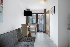 Una habitación con sofá, televisor montado en la pared, comedor con sillas y mesa, y puerta que da a un espacio exterior.
