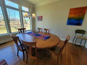 A dining area with a round wooden table, six chairs, a colorful placemat, a chandelier, and a large window with a view of trees and a house. - Cuthbert Place | Single-Level, 1.5mi Mayo, Pets OK (Rochester)