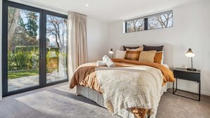 A bedroom with a large bed, a nightstand, a lamp, and a sliding glass door leading to a garden. - MacAndrew Luxury Lodge (Wānaka)