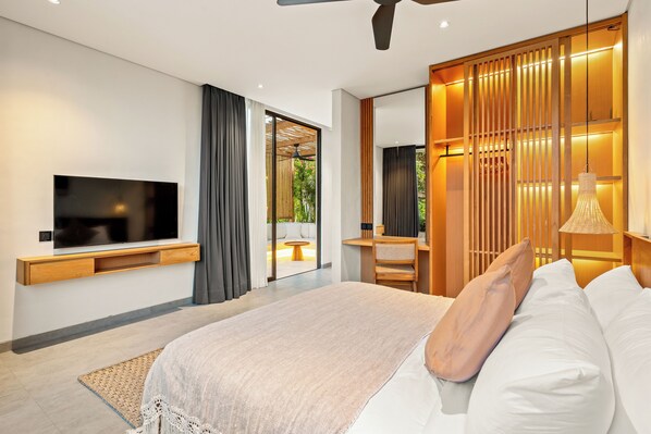 A modern bedroom with a large bed, a flat-screen TV mounted on the wall, a desk with a chair, and a sliding glass door leading to an outdoor area. - Villa Lago (Pecatu)