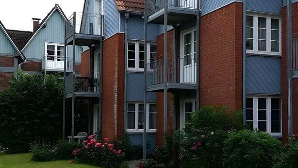 A multi-story residential building with balconies, brick and siding exteriors, and a well-maintained garden with flowers.