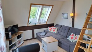 A loft-style room with a gray sofa, a small white side table, a black office chair, a wooden ladder, and a window with a view of greenery.