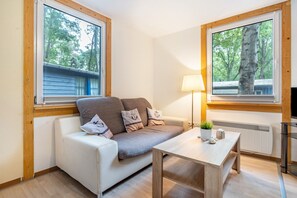 A room with a white sofa, a small table, a lamp, and two windows with views of trees and a building.