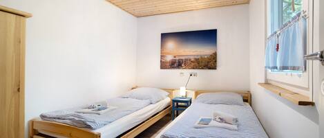 Een kamer met twee houten bedden, een nachtkastje met een lamp, een raam met gordijnen en een schilderij aan de muur.