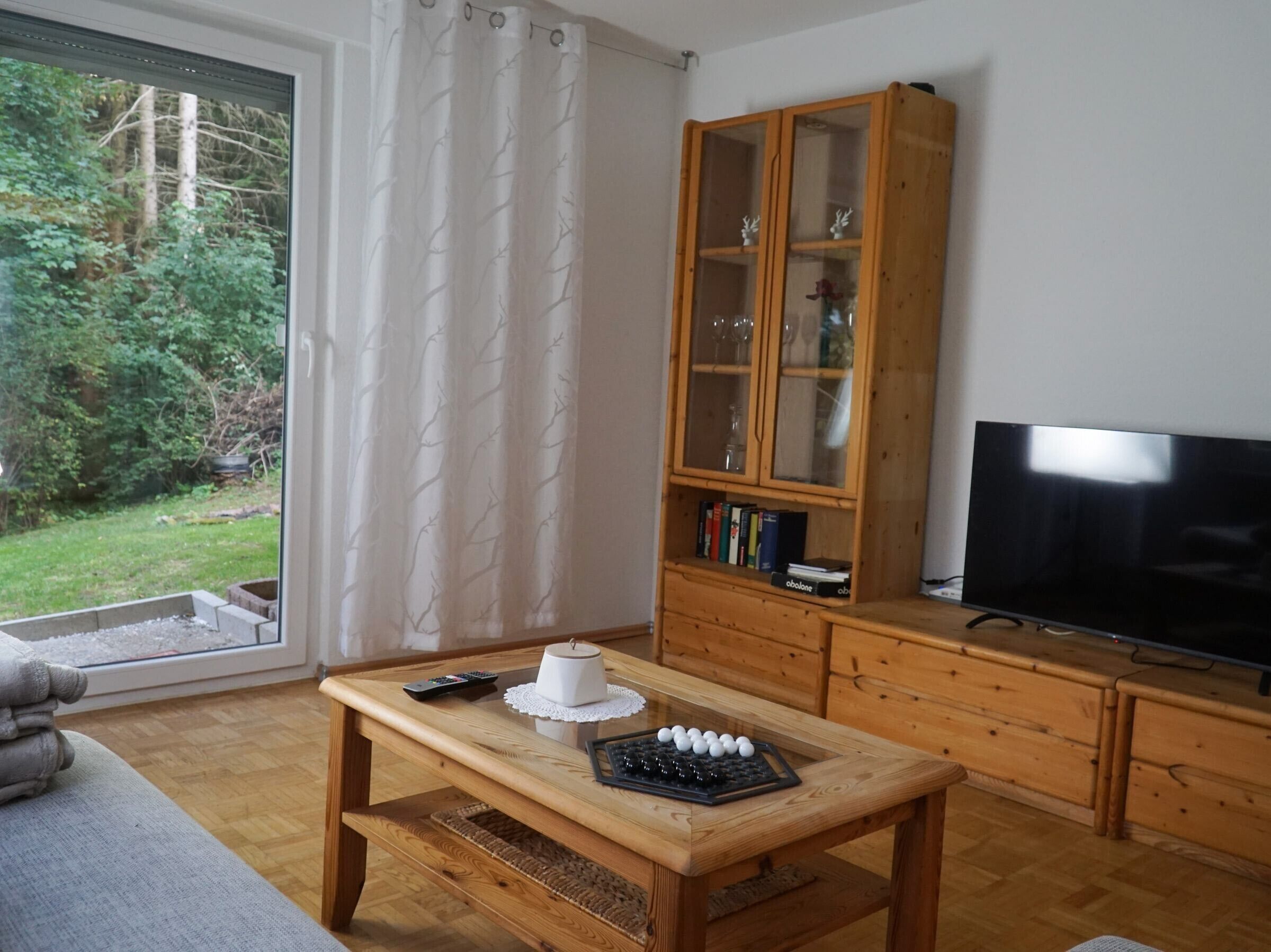 Una sala de estar con una mesa de madera, un mueble con puertas de vidrio, un televisor y vistas a un bosque por la ventana.