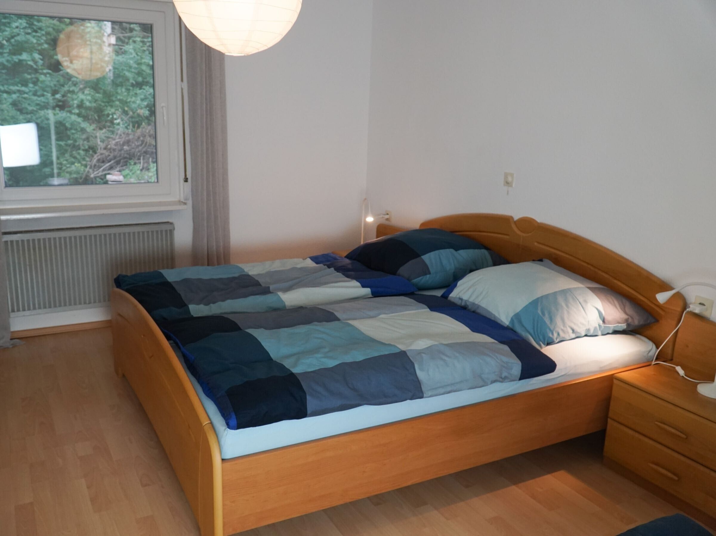 Een slaapkamer met een houten bed, nachtkastje, lamp, raam en een radiator.