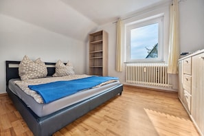 Un dormitorio con cama, suelo de madera, una ventana con cortinas, un radiador y un armario.