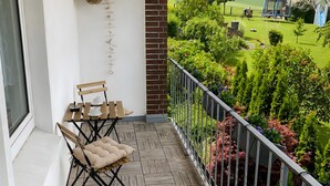 A balcony with a wooden floor, a table with chairs, a hanging plant, and a view of a green landscape.