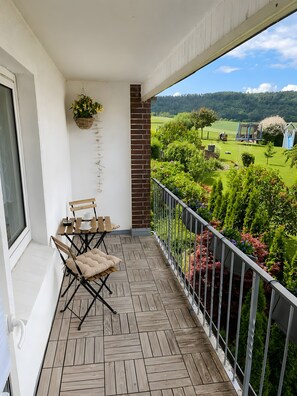 Un balcon doté d’un plancher en bois, une table avec des chaises, une plante suspendue et une vue sur un paysage verdoyant.