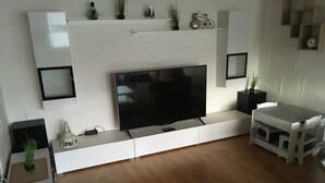 A modern living room with a large flat-screen TV, white cabinetry, a dining table, and a small bicycle on the wall. - Apartment 'Fewo Eberau' with Shared Garden and Wi-Fi (Ebrach)