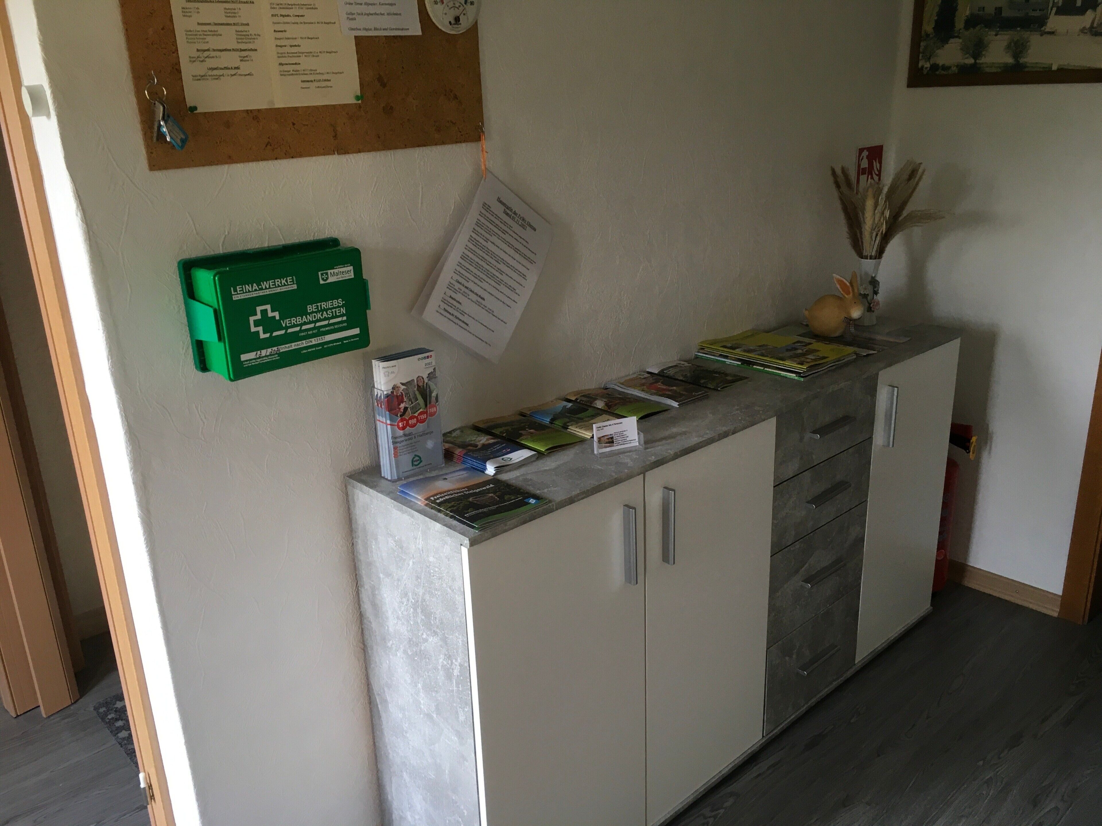 A reception desk with a corkboard, a green box labeled 'Leina-Werke,' and a small vase with dried plants.