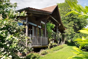 A wooden house with a porch, surrounded by lush greenery and flowering plants.
