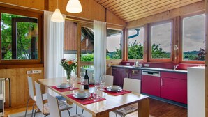 Un intérieur en bois avec une table à manger dressée pour quatre personnes, une cuisine aux armoires roses et de grandes fenêtres offrant une vue sur la verdure.