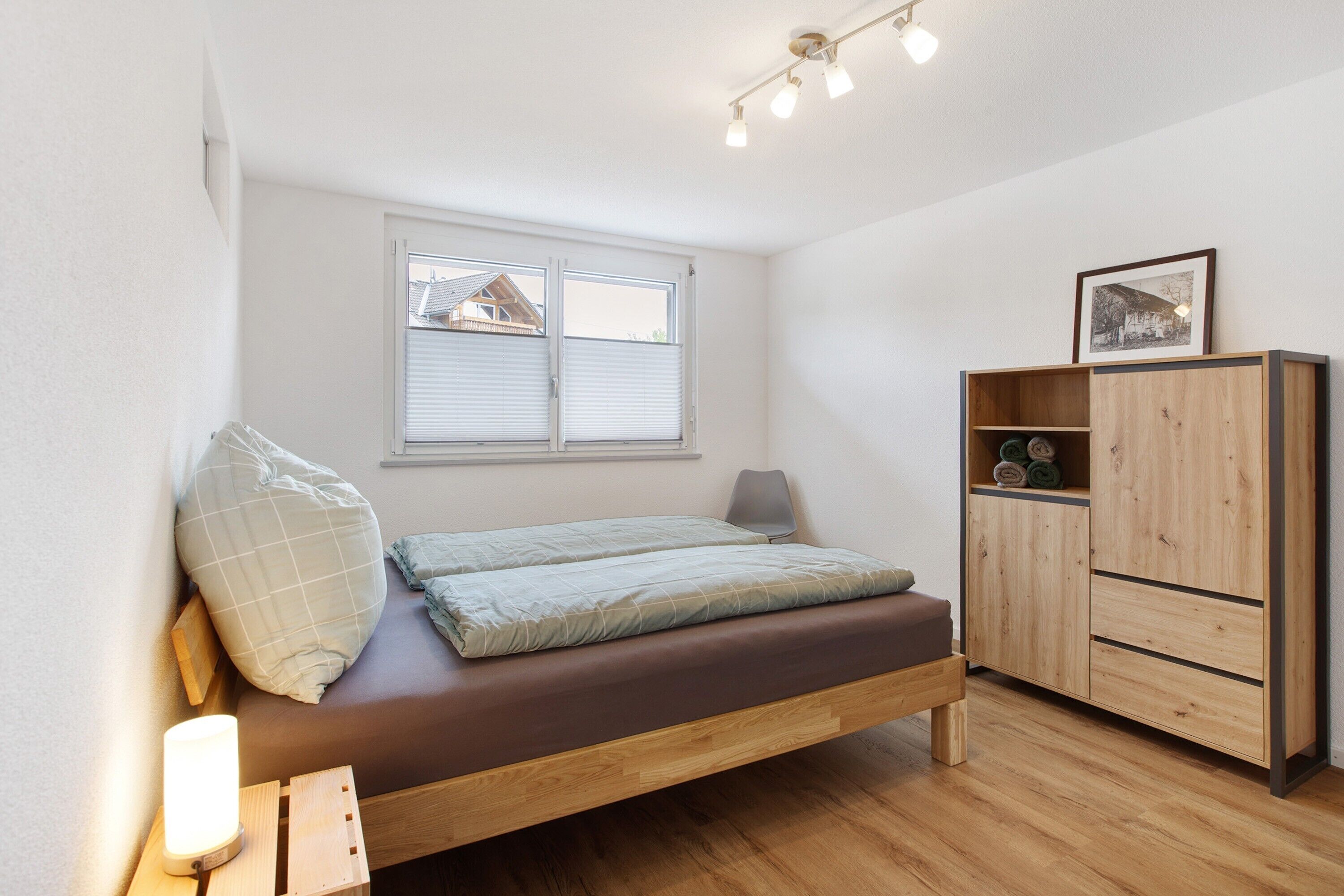A bedroom with a bed, a window, a bedside table with a lamp, and a wooden wardrobe.