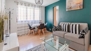 Una sala moderna con una mesa de centro de vidrio, un sofá con almohadones a rayas, una zona de comedor con mesa y sillas, y un ventanal con cortinas translúcidas.