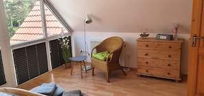 Une pièce avec une grande fenêtre, un fauteuil en osier avec un coussin, une petite table, une commode et un radiateur.