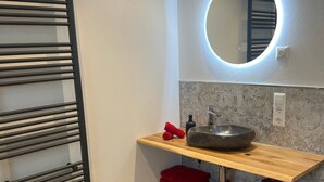 A bathroom with a towel rack, a round mirror with a light, a sink, and shelves with towels. - Holiday Home Ferienhaus am Kapellenhof with Shared Terrace, Shared Garden and Wi-Fi (Triberg im Schwarzwald)