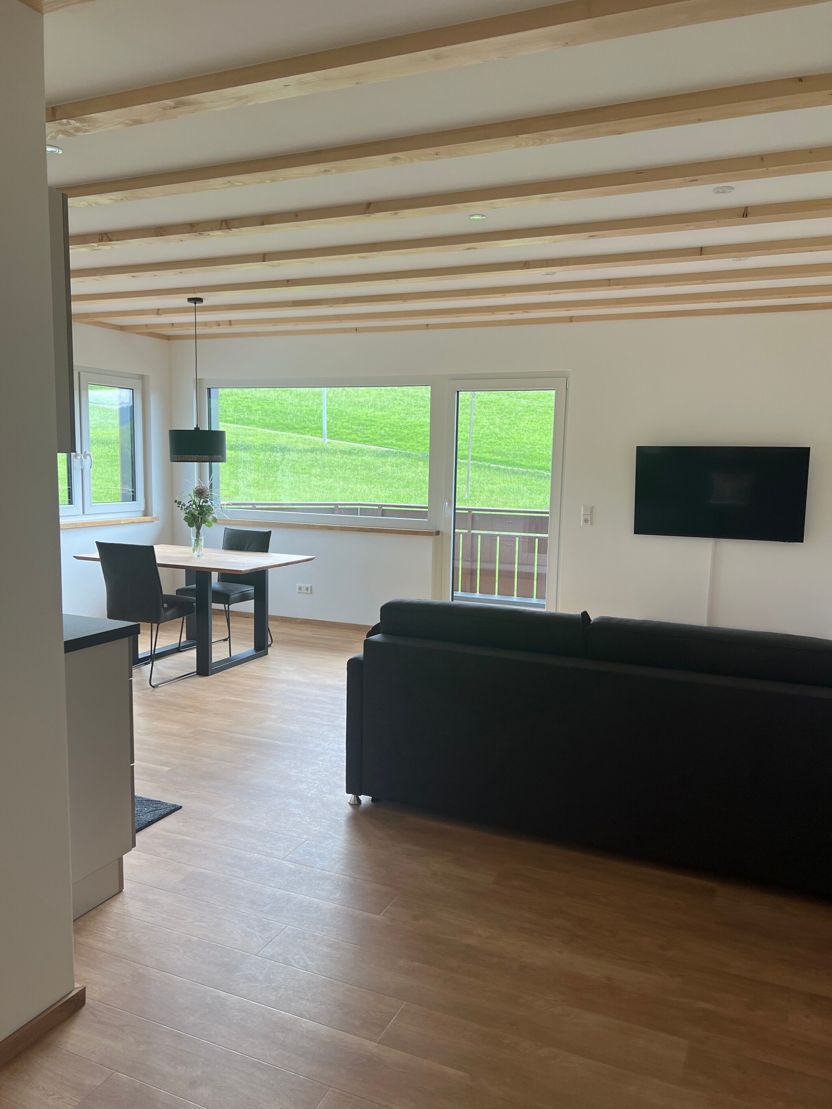 A spacious living area with a wooden ceiling, a black sofa, a dining table with chairs, and a TV mounted on the wall.