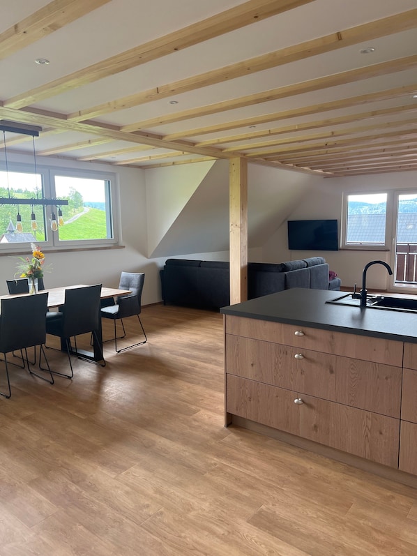 Une cuisine moderne avec des poutres en bois, un coin repas avec une table et des chaises, un canapé et une grande fenêtre donnant sur un paysage verdoyant.