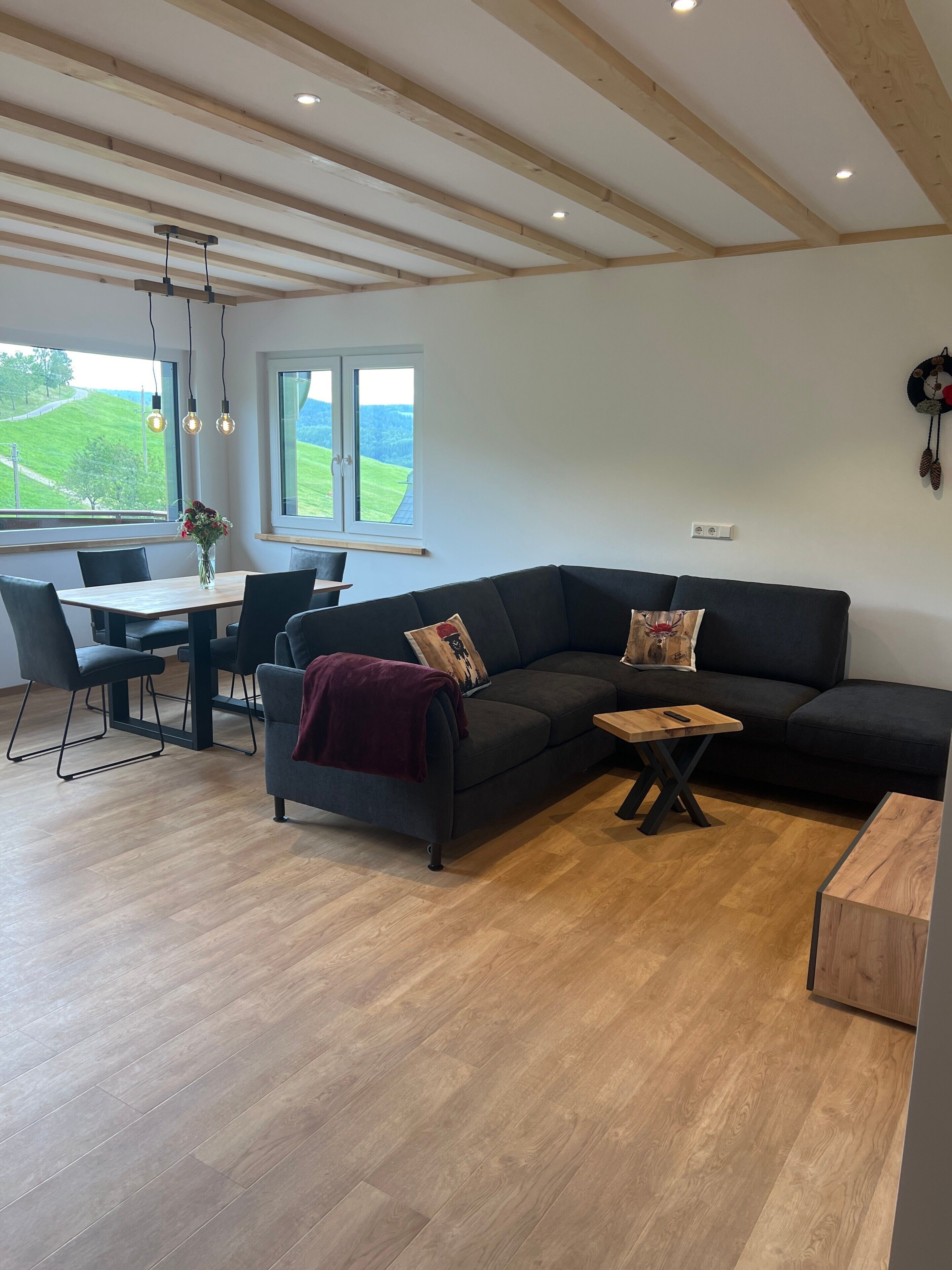 Un salón moderno con un sofá seccional gris oscuro, una mesa de centro de madera, una mesa de comedor con sillas y un ventanal que ofrece vistas a un paisaje verde.