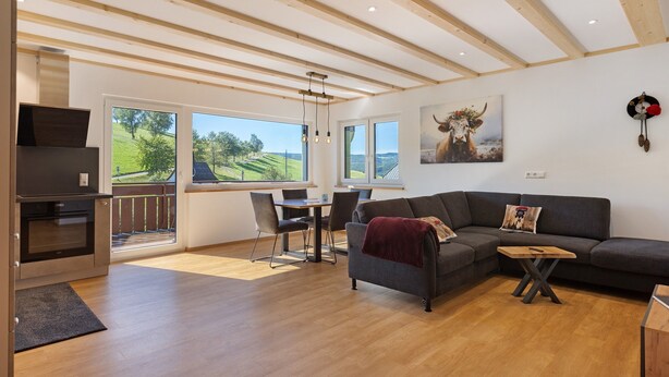 A modern living room with a wooden ceiling, a gray sofa, a dining table, and a kitchen area with an oven. - Apartment 'Kapellen Blick' with Mountain View, Shared Terrace and Wi-Fi (Triberg im Schwarzwald)