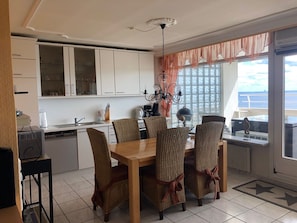 A dining area with a wooden table and wicker chairs, a kitchen with white cabinets, and a view of the sea through large windows. - Wellness Penthouse Sea View Apartment with Shared Garden and Wi-Fi (Sierksdorf)